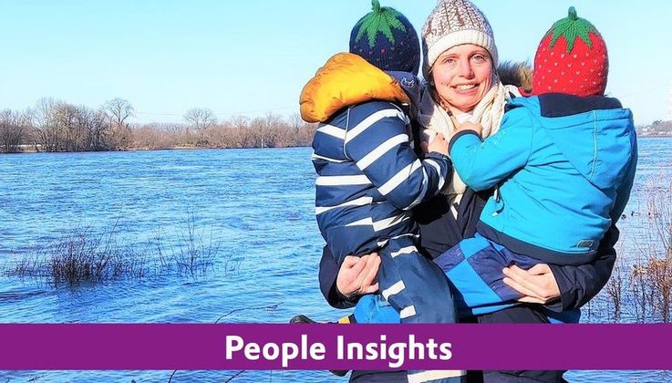 Eine Frau hat 2 Kinder auf dem Arm und posiert vor einem See.